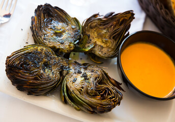 Halved artichokes cooked on charcoals served with romesco sauce on white plate. Catalan cuisine