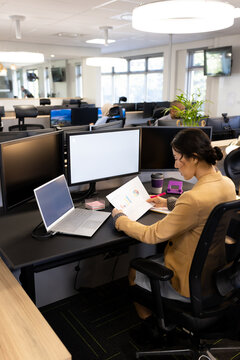 Vertical image of asian woman sitting at desk and working on computer with copy space in office