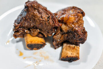 Roasted bone in beef short ribs cooked brown meat on white plate macro closeup with fat texture and...