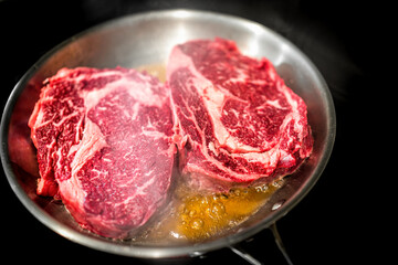 Stainless steel pan and closeup of two raw ribeye meat steaks cooking on stove in oil fat grease...