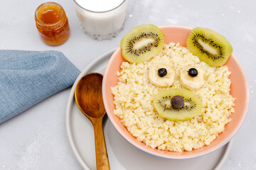 Funny cute kids childrens baby's healthy breakfast lunch oatmeal porridge in bowl look like bear,mouse face decorated with kiwi, banana, dried berry fruits,milk,honey. dessert food art on gray table