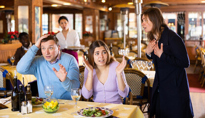 Resentful wife scolding her husband spending time with another woman at dinner in restaurant. Jealousy and betrayal concept.. - obrazy, fototapety, plakaty
