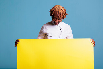 Professional cook wearing kitchen uniform showing a yellow banner on blue background in studio photo. Smiling afro american chef looking at placard available for copy space.