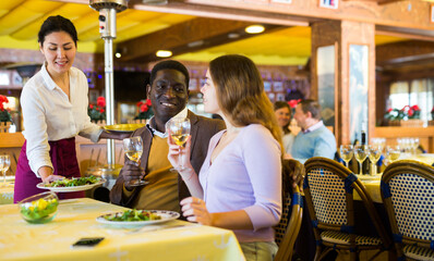 Caucasian woman and African-american man eating in restaurant during date. Asian woman waitress putting dish with salad on table.