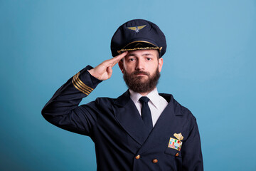 Obraz premium Serious aviation academy airplane pilot saluting, wearing uniform and hat front view portrait, plane captain looking at camera. Middle age aviator with airline wings badge on jacket