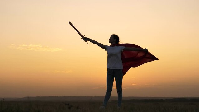Girl dreams of becoming superhero.Young girl plays superheroes. Child plays Spartan In. Free girl in red cloak with sword in her hand plays medieval knight, sun. Girl fights with toy sword. Childhood