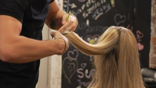 Male hairdresser cutting his female client's blonde hair with scissors and comb. Blurred blackboard in the background. High quality 4k footage