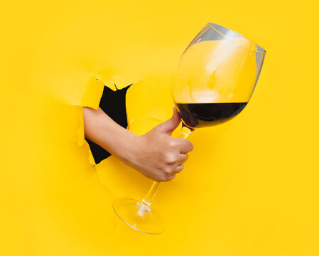 A man's hand emerges through a torn hole in yellow paper with a large glass of red wine. The concept of alcoholism, drunkenness and hangover. Like thumb and good quality alcohol.