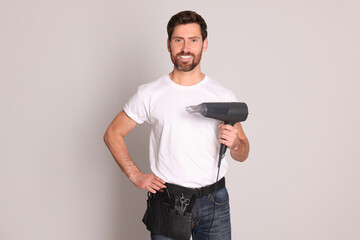 Smiling hairdresser with tool bag holding dryer on light grey background