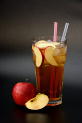 Apple juice in a tall glass with ice and straws on a black background, next to it are red ripe fruits.