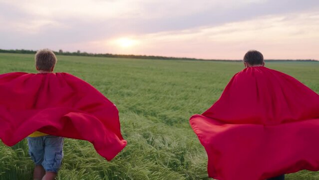 Happy boys play superheroes they run across green field in red cloak, cloak flutters in wind. Childrens games and dreams. Slow motion. teenager dreams of becoming superhero. Young boys in red cloak