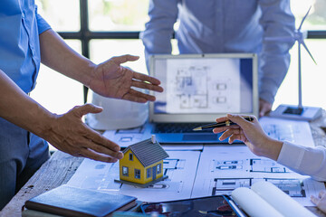 Male engineer meeting pointing at blueprint with house plan and wind turbine next to house building...