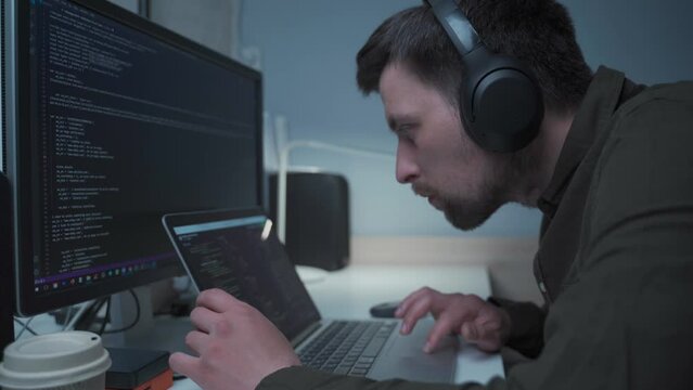 Handsome male software developer programming codes while working behind laptop with headphones from home. IT engineer in headphones working on computer in office. Freelancer works, social distancing.