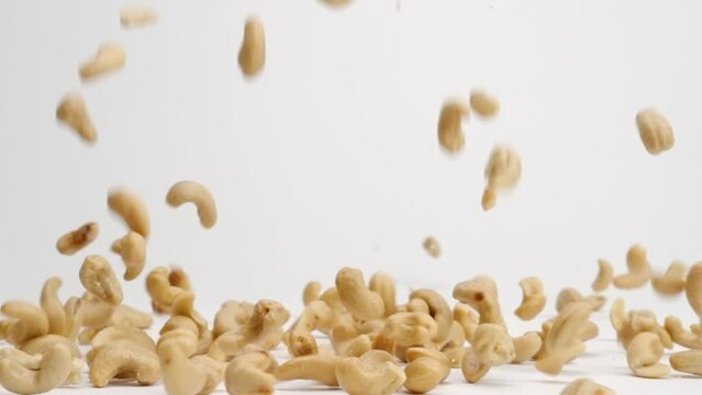 Whole roasted and salted cashew nuts falling onto white table top and bouncing around before landing in a pile in slow motion