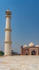 Obraz premium The tall white marble minaret of the ancient mausoleum of the Taj Mahal against the blue sky. Behind you can see a red sandstone Kau Ban mosque with domes and arches. India. Agra