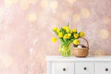 Basket with Easter eggs and vase with yellow tulips on table near grunge wall