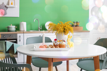 Tray with Easter cakes, eggs, burning candle, bunny and mimosa flowers on dining table in kitchen