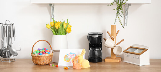 Basket with Easter candies, rabbits, greeting card and vase with tulips on counter near light wall in kitchen