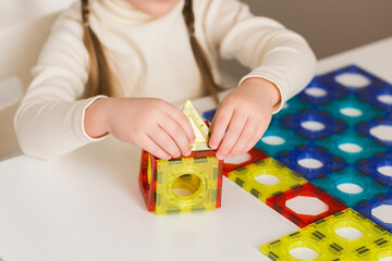 A child plays with a magnetic constructor on a white table at home or in kindergarten. Building a toy house from blocks. Hands close up.