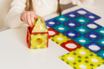 A child plays with a magnetic constructor on a white table at home or in kindergarten. Building a toy house from blocks. Hands close up.