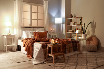 Interior of bedroom with brown checkered blankets on bed, burning candles and glowing lamps late in evening