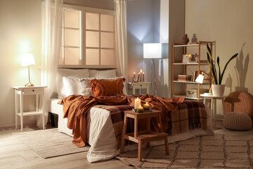 Interior of bedroom with brown checkered blankets on bed, burning candles and glowing lamps late in evening