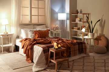 Interior of bedroom with brown checkered blankets on bed, burning candles and glowing lamps late in evening