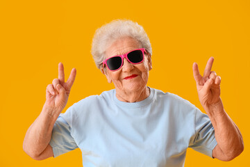 Naklejka premium Senior woman in sunglasses showing victory gesture on yellow background