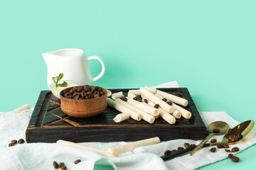 Black wooden board with delicious wafer rolls and bowl of coffee beans on turquoise background