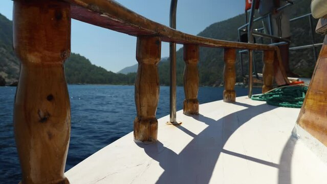 White wooden deck of a pleasure boat with railings and balusters. In the background is the sea, mountains and forest. The tourist boat is heading towards the shore. A summer holiday