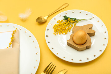 Table setting with Easter eggs, mimosa flowers and golden cutlery on yellow background