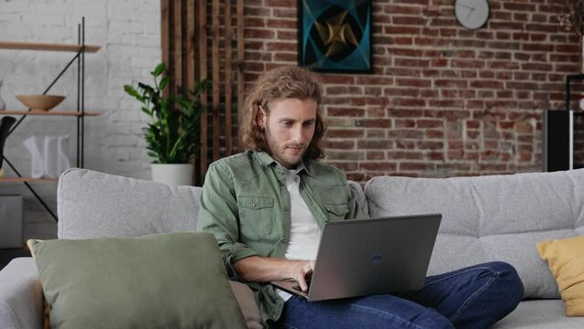Relaxed Man user holding laptop computer sit on sofa at home. Focused male using notebook check online news. Focused curly Guy texting Messages on his Laptop in living room at home. Indoor. Freelance.