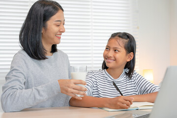 Asian young little girl learn at home. Do homework with kind mother help, encourage for exam. Mom pass on a glass of milk to daughter. Girl happy Homeschool. Mom teach and advice education together.