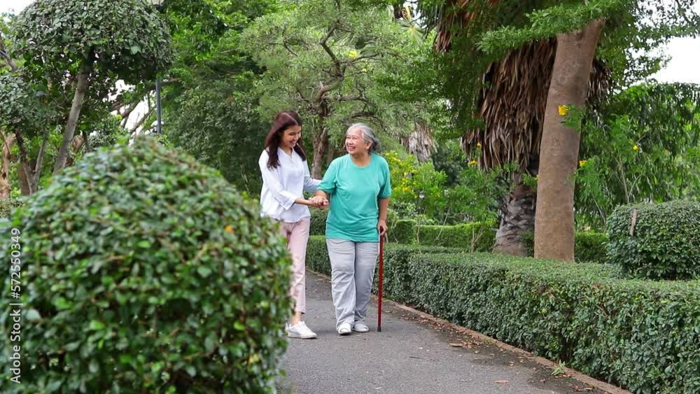 Wall mural A caregiver helps support an elderly Asian woman holding a walking cane for exercise in a park. Elderly health care. senior physiotherapy