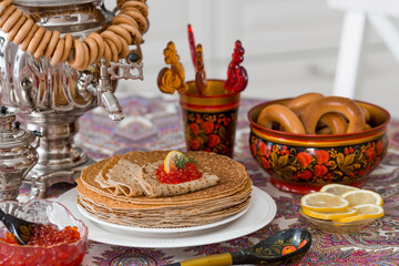 Russian traditions. Russian holiday Maslenitsa. Still life with a cup of tea, a stack of pancakes, red caviar, lemon, bagels, a lollipop cockerel, wooden spoons (khokhloma) and samovars.