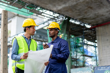 African American Technician, Asian Engineer use blueprint to inspect construction. Group of Multi-Ethnic Blue-collar worker. Teamwork of Architect review plans at construction site.