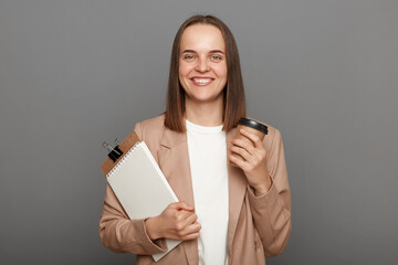 Image of smiling happy woman with brown hair wearing beige jacket, holding organizer and drinking coffee to go, looking at camera with positive expression, posing isolated over gray background.