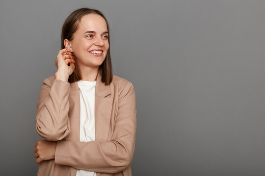 Image of attractive delighted woman with brown hair wearing beige jacket, smiling while looking aside at mockup for advertisement, posing isolated over gray background.