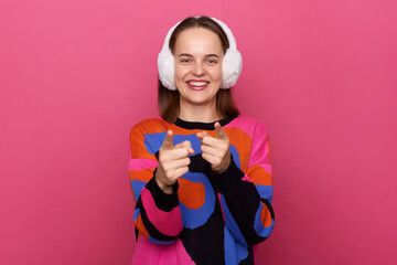 Indoor shot of smiling cheerful woman wearing sweater and earmuffs, pointing to camera, showing direction and choosing, indicating with index finger, posing isolated over pink background.