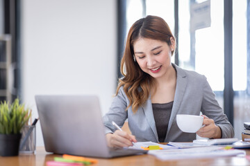 Business Asian woman using calculator and writing make note with calculate doing math finance on an office desk. Woman working at office with laptop and tax, accounting, documents on desk.