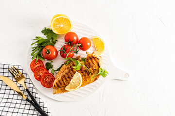 Grilled chicken breast in different variations with cherry tomatoes, green herbs, cut lemon on a white ceramic serving tray. Top view.