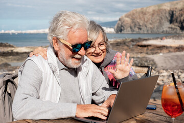 Smiling senior couple in video chat with laptop enjoying sunny day outdoor at the beach sitting at bar wooden table with orange cocktail. Elderly people in vacation or retirement at sea