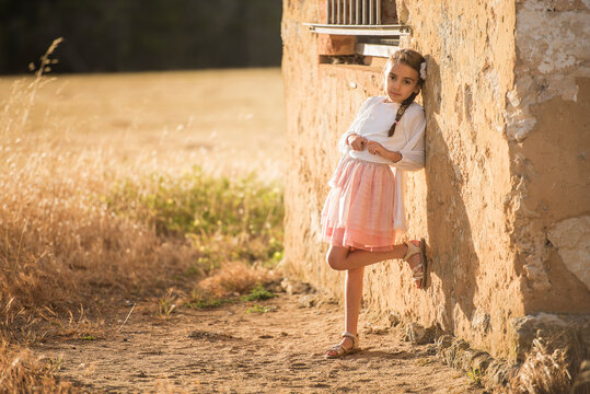 little girl in pink skirt leaning against rustic wall at golden hour
