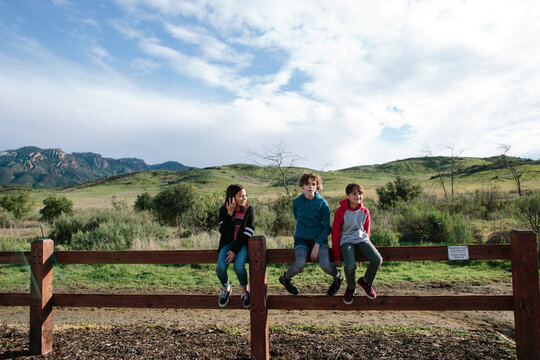 Three siblings sit on a wood fence outside near a nature trail