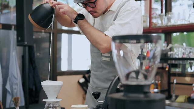 Asian man barista wearing an apron uses a tea kettle to make pour over coffee at a cafe during the day&nbsp;