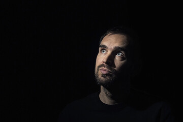 A portrait of a man with a small beard staring thoughtfully upwards to the side. Close-up. A middle-aged man against a black background wearing a black T-shirt. Light illuminates half of his face.