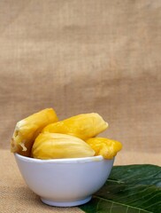 Ripe Jackfruit Pieces in a White Bowl with Green Leaves Isolate on Burlap Fabric with Copy Space in Vertical Orientation