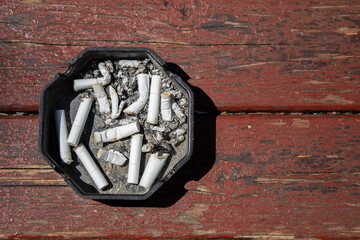 ashtray full of cigarette butts on brown wooden table, top view in daylight