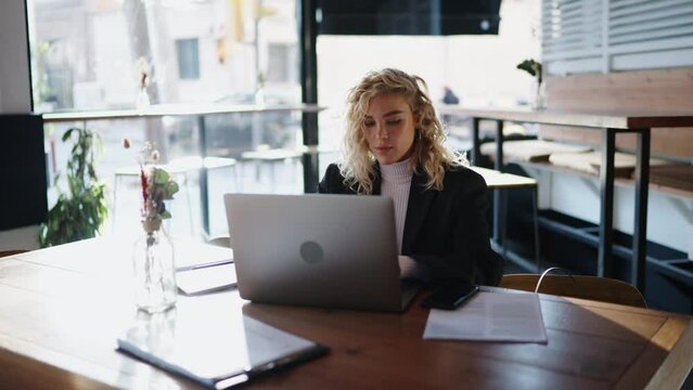Concentrated female using laptop computer while sitting in urban cafe. Young attractive woman administrator keeps records in electronic form and solves the work tasks of coffee shop. Slow motion