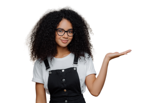 Horizontal shot of pretty dark skinned curly girl raises palm, holds empty space, wears transparent glasses, white t shirt and overalls, isolated over brown background, advertises some object